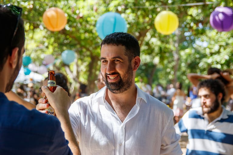 גבר בחולצה לבנה מחייך ומחזיק משקה, פנסי נייר צבעוניים מעליו