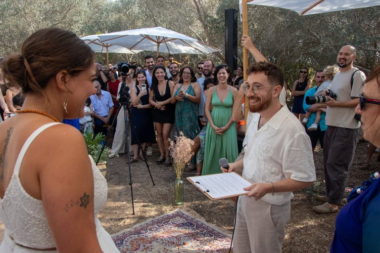 גבר בחולצה בהירה מחזיק דפים ומדבר מול אורחים, והכלה בקדמת התמונה