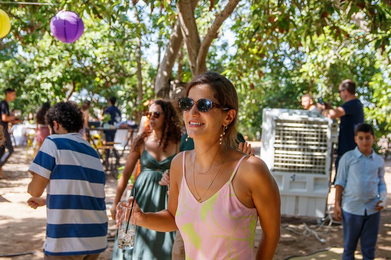 אישה במשקפי שמש מחייכת, אורחים מאחוריה בין העצים