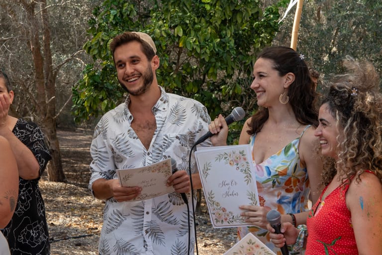 גבר ואישה נואמים ליד מיקרופון ומחזיקים כרטיסים מעוטרים, אורחים לצידם