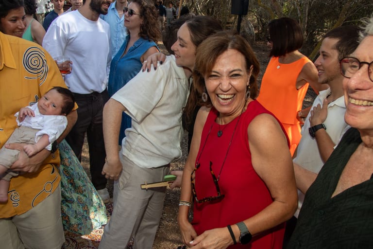 אישה מחייכת בשמלה אדומה מחבקת אורחים, ותינוק בזרועות אחד מהם