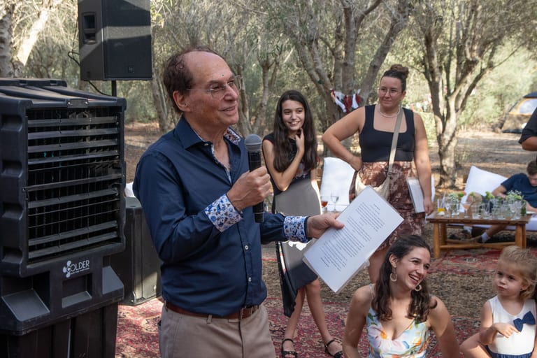 גבר מבוגר בחולצה כחולה קורא נאום אל תוך מיקרופון מול אורחים