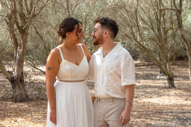 הכלה והחתן עומדים פנים אל פנים בחיוך בחורשת זיתים