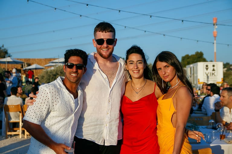 ארבעה אורחים מחייכים יחד בשעת השקיעה, שתיים מהן בשמלות אדומה וכתומה