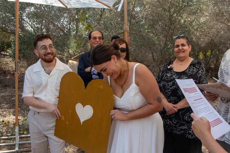 הכלה מסתירה את פניה בצחוק מאחורי שלט זהב בצורת לב, והחתן לצידה