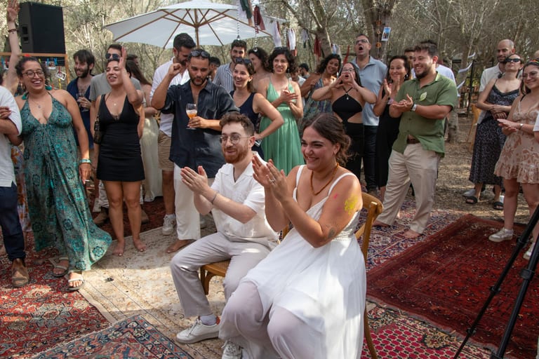 הכלה והחתן יושבים על כיסאות והאורחים מוחאים כפיים סביבם
